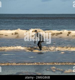 Surfeur mâle caucasien portant une combinaison noire debout sur une planche de surf arrivant sur terre à Cayton Bay, North Yorkshire, Royaume-Uni Banque D'Images