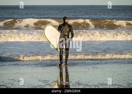 Surfeur mâle caucasien portant une combinaison noire émergeant de la mer transportant une planche à voile à Cayton Bay, North Yorkshire, Royaume-Uni Banque D'Images