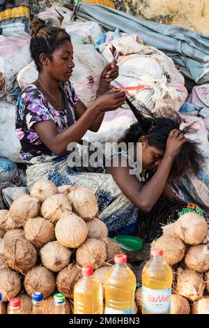 Deux femmes braiding cheveux, marché décrochage, noix de coco, sacs de charbon derrière, Mahajanga, Madagascar Banque D'Images