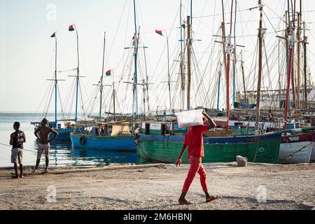 Homme en vêtements rouges portant sac, bateaux de pêche derrière, vieux port, Mahajanga, Madagascar Banque D'Images