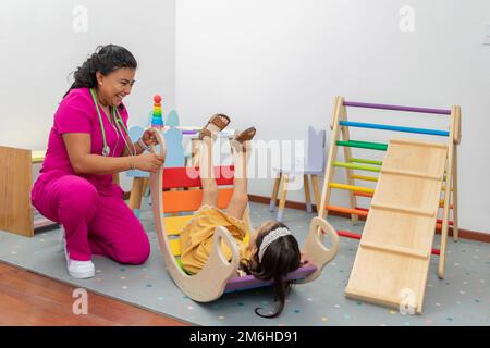 Latina médecin pédiatrique, riant en jouant avec une fille dans la salle de jeux de son bureau . Banque D'Images