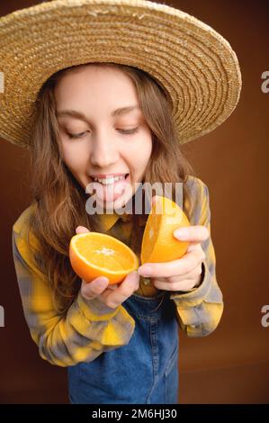 Portrait studio d'une jeune femme caucasienne attrayante dans un chapeau et une combinaison en denim tient un fruit orange coupé dans ses mains et sho Banque D'Images