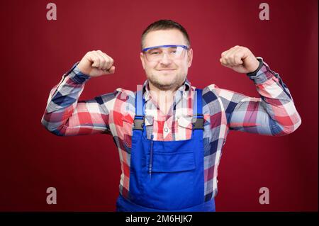 Portrait d'un réparateur de contremaître homme caucasien amical positif dans des combinaisons et des lunettes sur fond rouge foncé. Looki souriant Banque D'Images