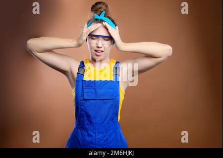 Jeune femme caucasienne attirante travailleuse de construction dans l'uniforme d'un handyman tient sa tête avec ses mains de a Banque D'Images