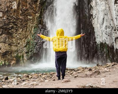 Une femme dans une veste jaune se tient avec ses mains à part, regardant une cascade de glace de tempête coulant sur des rochers humides. Vue arrière Banque D'Images