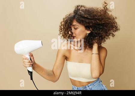 Femme sèche les cheveux freux avec sèche-cheveux, maison beauté produits coiffants cheveux, sourire sur fond beige Banque D'Images