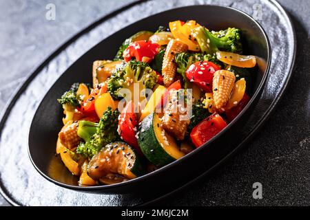 sauté de légumes de poivron, oignon, courgettes, maïs en petits épis, bouquets de brocoli versés avec de la sauce soya collante dans un bol noir , gros plan Banque D'Images