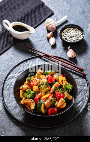 sauté de légumes de poivron, d'oignon, de courgettes, de maïs en rafles, de fleurons de brocoli versés avec de la sauce soya collante dans un bol noir avec des baguettes, et de gousse Banque D'Images