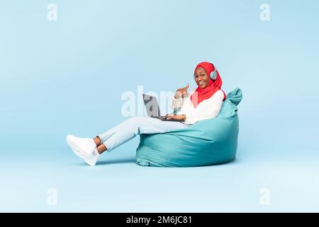 Bonne femme musulmane noire dans un casque, assise sur une chaise avec un ordinateur portable et montrant le pouce vers le haut, fond bleu Banque D'Images
