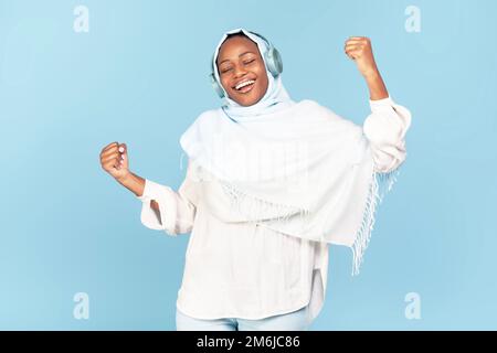 Joyeuse femme noire dans hijab écoutant de la musique en ligne dans des écouteurs sans fil, appréciant la chanson préférée, fond bleu Banque D'Images