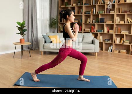Yoga domestique. Vue latérale d'une jeune femme japonaise qui fait une fente, tient les mains dans le namaste, s'exerçant à la maison, pleine longueur Banque D'Images