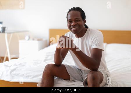 Jeune homme noir assis sur le lit dans la chambre et souriant à la caméra Banque D'Images