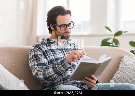 Sérieux élégant jeune caucasien étudiant en lunettes et chemise est assis sur le canapé, lit le livre, profiter de temps libre Banque D'Images