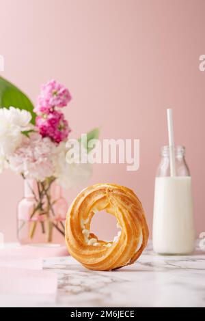 Profiterole circulaire maison avec sucre en poudre couvert de crème anglaise sur table en marbre sur fond rose avec fleurs et lait Banque D'Images