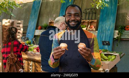 Jeune vendeur masculin présentant des produits biologiques naturels et parlant à des clients sur le marché des agriculteurs. Marché à l'extérieur avec fruits et légumes biologiques cultivés localement, porte-stalle de marché. Banque D'Images