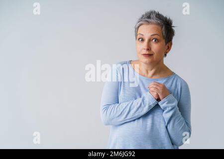 Une femme mûre stupéfait en 50s avec des cheveux gris posant avec les mains à côté du cœur et l'espace de copie sur la gauche isolée sur le backgroun blanc Banque D'Images