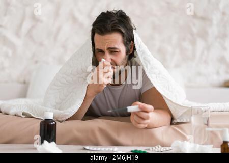 Jeune caucasien triste et découplé malade mâle se trouve sur le lit sous une couverture, souffre d'écoulement, souffle son nez dans la serviette Banque D'Images