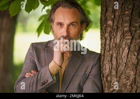 Charmant homme d'affaires mature et bibord gris décontracté debout sous l'arbre sur l'herbe regardant la caméra avec les bras repliés touchant h Banque D'Images