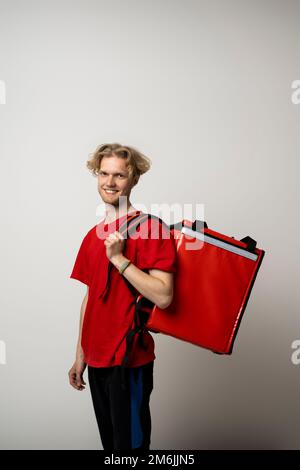 Jeune coursier souriant avec cheveux bouclés en uniforme rouge et sac Thermo rouge sur fond blanc. Service de livraison de nourriture. Banque D'Images