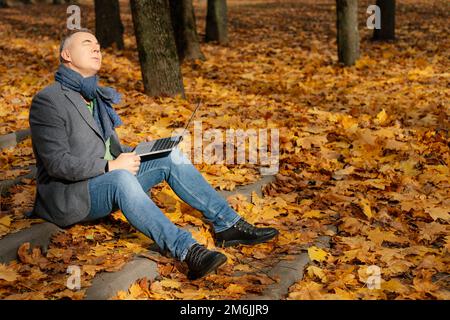 Vue latérale d'un homme d'âge moyen assis avec les yeux fermés sur les escaliers recouverts de feuilles jaunes en automne, tenant un ordinateur portable. Banque D'Images