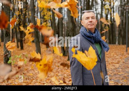 Portrait d'un beau homme d'âge moyen debout près des arbres parmi les feuilles mortes jaunes volantes dans la forêt du parc en automne. Banque D'Images