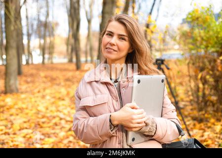 Portrait d'une femme d'âge moyen debout près d'arbres sur des feuilles d'érable jaune tombées dans la forêt du parc en automne tenant un ordinateur portable. Banque D'Images