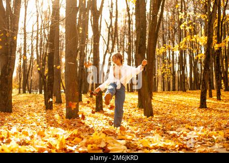 Portrait d'une jeune adolescente ludique marchant parmi les arbres dans la forêt du parc en automne, donnant des coups de pied aux feuilles d'érable jaune tombé. Banque D'Images