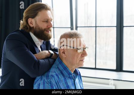 Le fils dans un costume élégant s'appuie sur les épaules de son père, le Père et le fils dans un arrière-plan énorme fenêtre, les deux hommes regardent dans le Banque D'Images