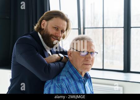 Le fils dans un costume élégant s'appuie sur les épaules de son père, le Père et le fils dans un arrière-plan énorme fenêtre, les deux hommes regardent dans le Banque D'Images