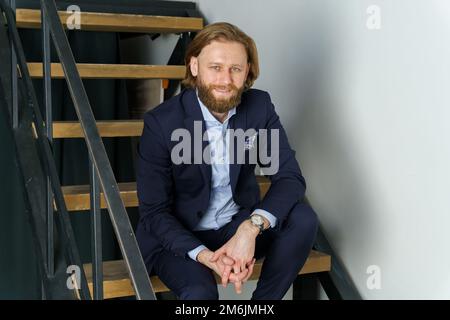 Un bel homme brutal barbu et aux cheveux longs est assis sur les marches de l'escalier, un homme d'affaires élégant Banque D'Images