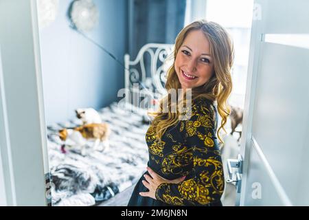 Prise de vue au niveau des épaules d'une femme blonde avec ses mains sur les hanches, regardant l'acmr et souriant. Devon rex jouant sur le lit en arrière-plan. Arrière-plan flou. Photo de haute qualité Banque D'Images