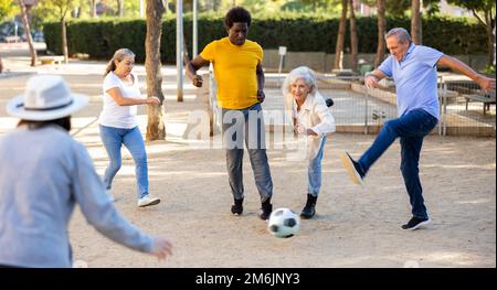 Joyeux amis multinationaux jouant au football sur un terrain de jeu sablonneux lors d'une chaude soirée de printemps Banque D'Images