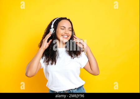 Joyeuse jolie brunette femme hispanique, dans un vêtement de base décontracté, avec des écouteurs, écoute sa musique préférée, danse avec les yeux fermés, se détendre, s'amuser sur fond jaune isolé, sourires Banque D'Images