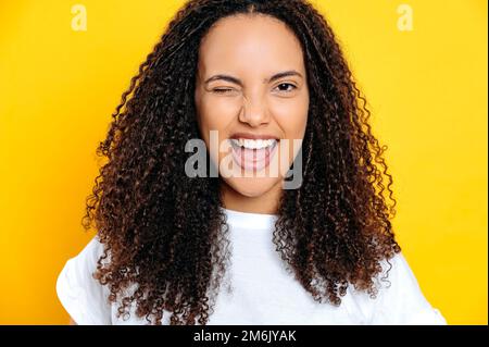 Photo en gros plan d'une charmante femme brésilienne ou hispanique joyeuse et joyeuse, vêtue d'un t-shirt blanc, envelouillée et souriante à l'appareil photo, debout sur un arrière-plan orange isolé Banque D'Images