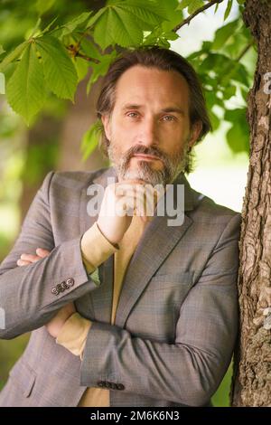 Homme d'affaires mûr et élégant, bibord gris, debout sous l'arbre sur l'herbe, regardant l'appareil-photo avec les bras repliés en touchant h Banque D'Images