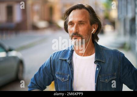 Beau homme mûr et mûr à barbe grise avec écouteurs qui écoute de la musique ou qui parle au téléphone, debout dans une rue urbaine Banque D'Images