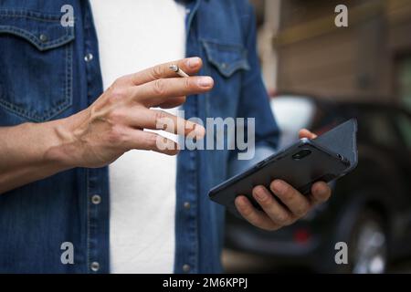 Fumer un cigaret homme d'âge moyen vérifiant des messages texte ou des médias sociaux à l'aide de son smartphone portant un Jean et un t-shir blanc Banque D'Images