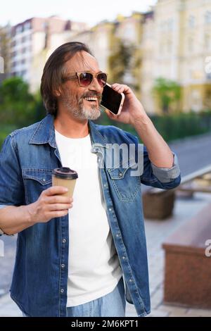 Homme mûr et enthousiaste avec une barbe grise parlant au téléphone tout en prenant un café en papier jetable tasse de travail ou de voyage debout Banque D'Images