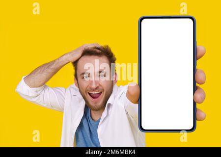 Beau jeune homme excité souriant émotionnellement touchant sa tête montrant un immense smartphone avec écran blanc. Publicités pour applications mobiles Banque D'Images