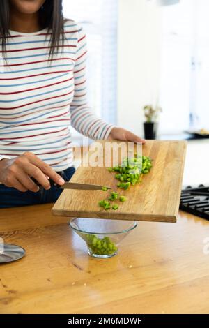 Section médiane de la jeune femme biraciale tenant une planche avec des poivrons verts hachés sur l'île de cuisine Banque D'Images