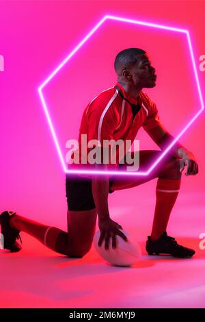 Un homme afro-américain avec un ballon de rugby agenouillé par un hexagone lumineux sur fond rose Banque D'Images