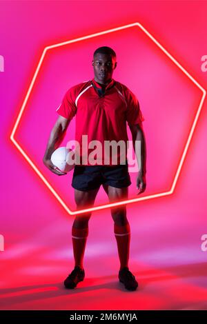 Joueur de rugby afro-américain plein de confiance avec un ballon sur un hexagone éclairé sur fond rose Banque D'Images