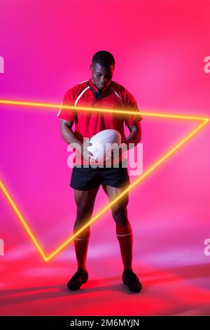 Joueur de rugby afro-américain plein de confiance avec balle par triangle lumineux sur fond rose Banque D'Images