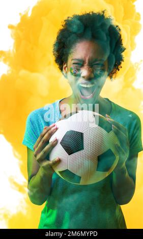 Fan de l'amérique africaine avec la peinture du visage et le ballon du drapeau brésilien criant sur fond fumé Banque D'Images