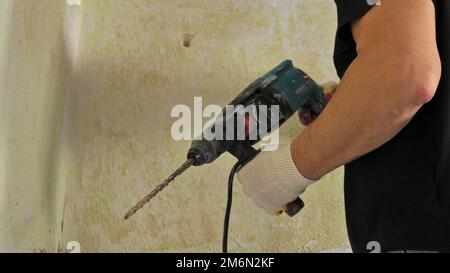 un homme en vêtements noirs tient un perforateur avec une longue perceuse et se tient près d'un mur de merde texturé, le travail de perçage de trous avec un outil puissant Banque D'Images