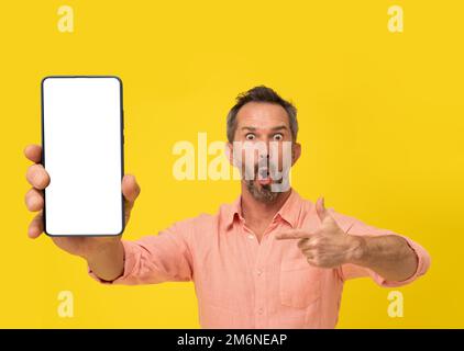 Émerveillé gris d'âge moyen homme à cheveux pointant le doigt vers un smartphone regardant l'appareil photo portant une chemise pêche isolée sur jaune. Matur Banque D'Images