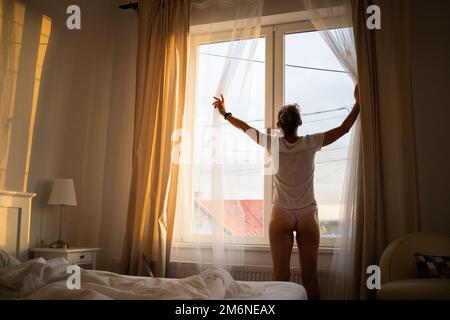 Femme ouvrant les rideaux de la chambre au lever du soleil Banque D'Images