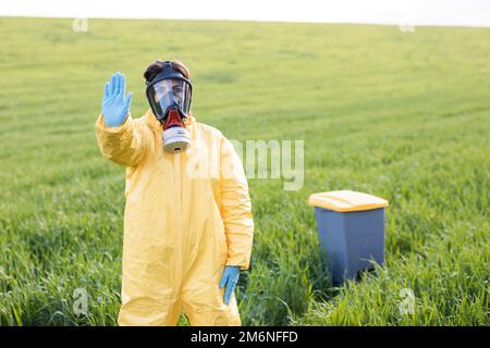 Une femme en costume de protection jaune se tient au milieu d'un champ vert dans un masque à gaz de protection et montre un geste d'appel pour arrêter et sauver le pl Banque D'Images