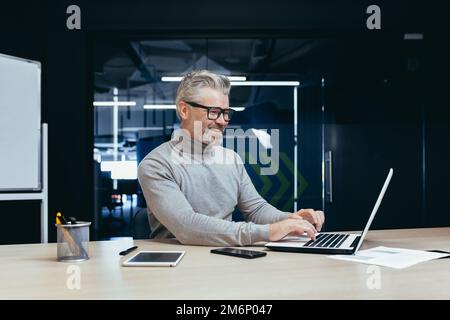Joyeux et souriant homme d'affaires mature travaillant à l'intérieur du bureau, homme aux cheveux gris senior utilisant un ordinateur portable sur le lieu de travail souriant satisfait avec de bons résultats. Banque D'Images
