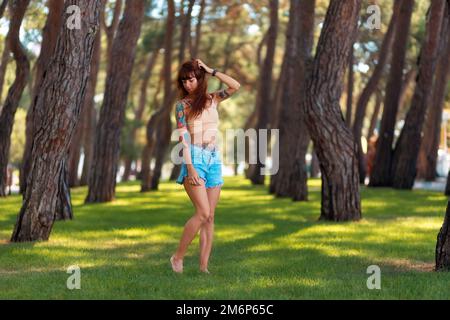 Caucasien mince belle jeune femme bronzée avec des tatouages posant des stands sur la pelouse dans un parc bosquet. Vacances d'été en plein air. Concept de psychologie Banque D'Images